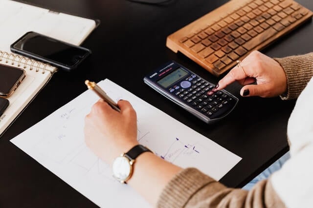 A person uses a calculator with one hand while writing on a sheet of paper with the other. A keyboard, notebook, and smartphone are on the desk.