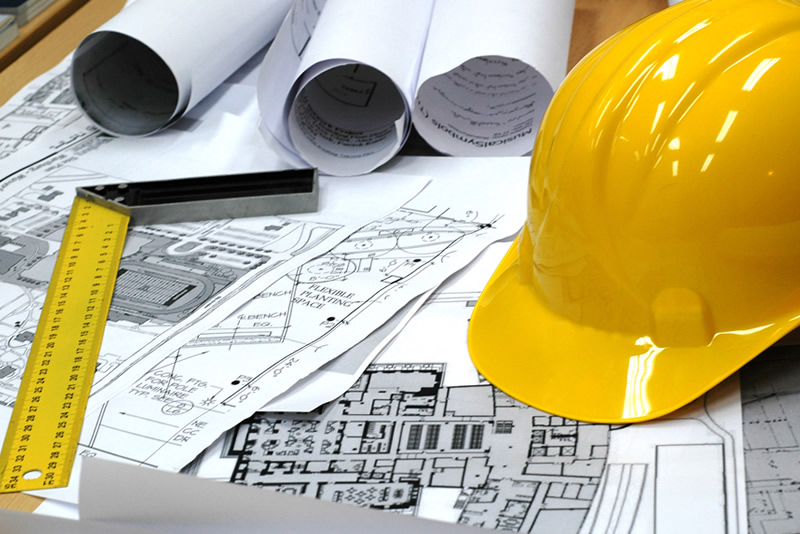 A yellow hard hat, measuring tools, and rolled-up blueprints are placed on top of architectural plans spread out on a desk, suggesting a construction or engineering project in progress.