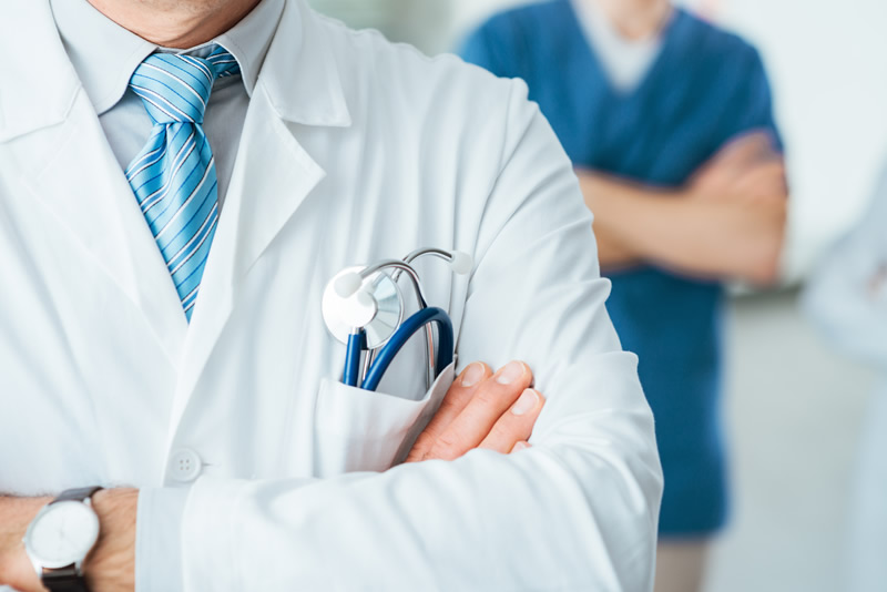A doctor in a white coat with a stethoscope in the pocket stands with arms crossed; another medical professional in blue scrubs is blurred in the background.